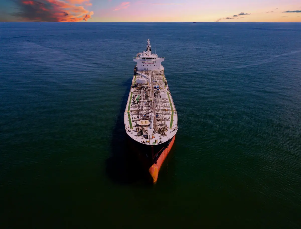 Aerial view of a crude oil tanker sailing on calm open water at sunset, its long deck of pipes running toward the bow