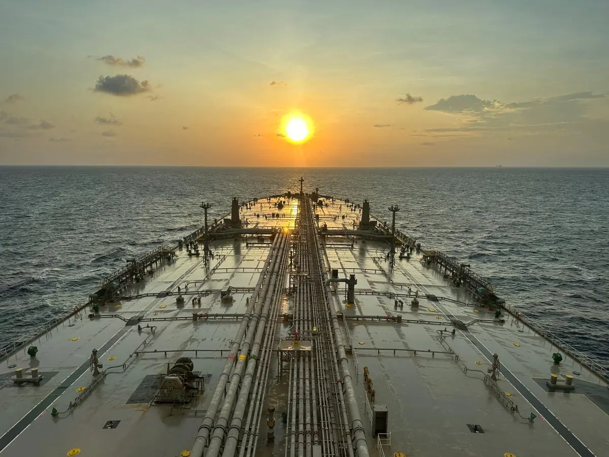 View along the deck of a crude oil tanker at sea, with pipelines running towards the bow and the sun setting on the horizon